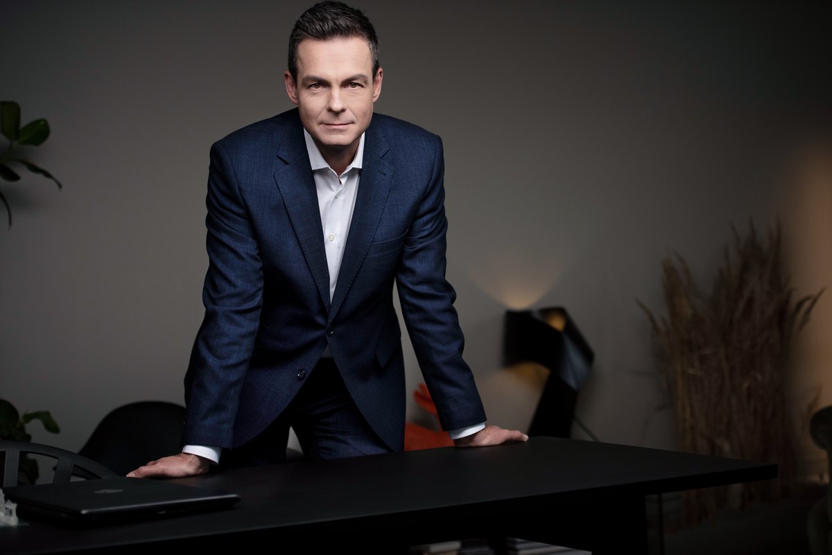 Mann im blauen Anzug stützt sich auf einen dunklen Tisch