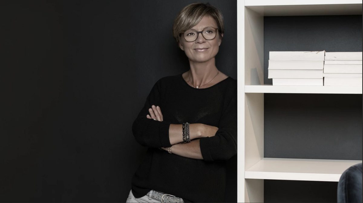 Frau mit Brille und verschränkten Armen lehnt neben einem weißen Bücherregal an dunkler Wand