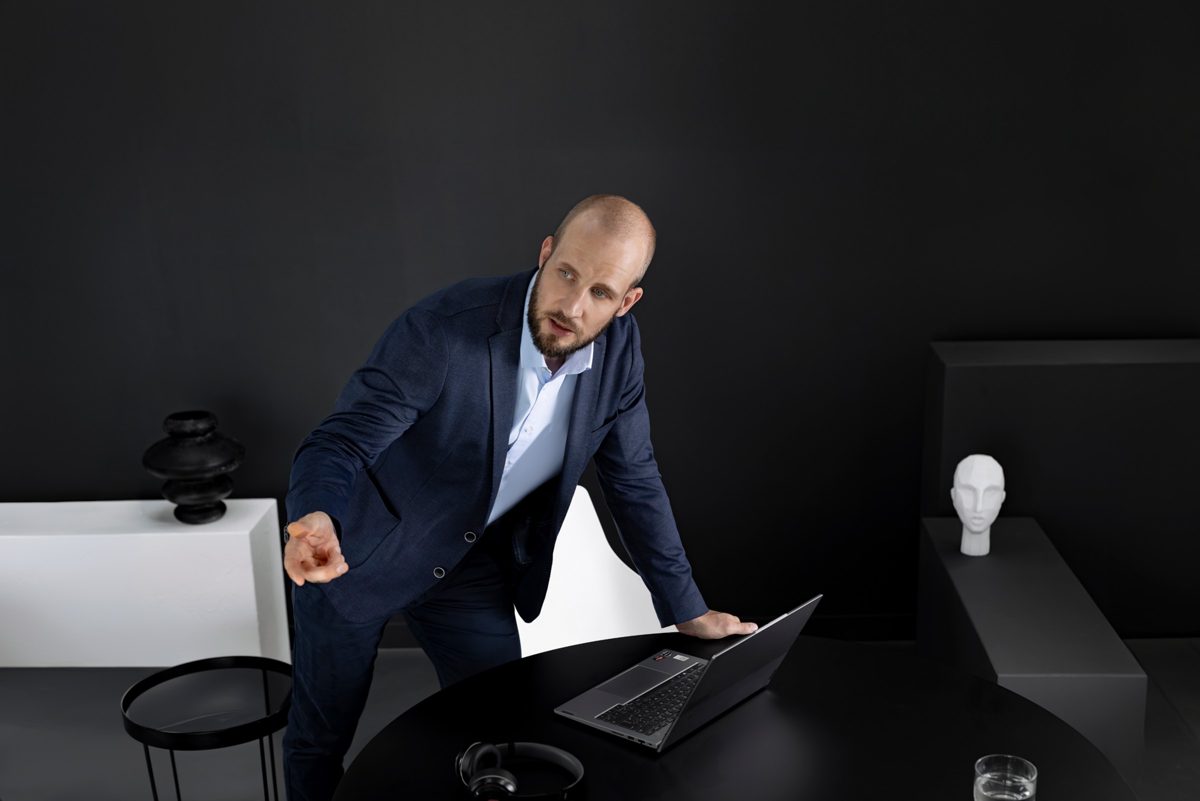 Mann im blauen Sakko gestikuliert über einem Tisch mit Laptop in schwarzer Studiokulisse