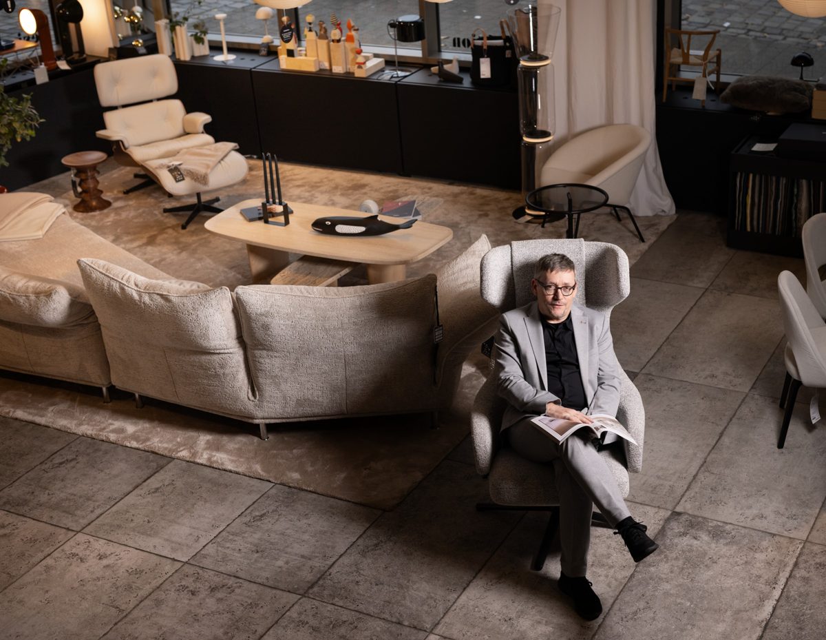 Mann im hellen Anzug sitzt mit Magazin in einem Sessel inmitten eines Möbel-Showrooms