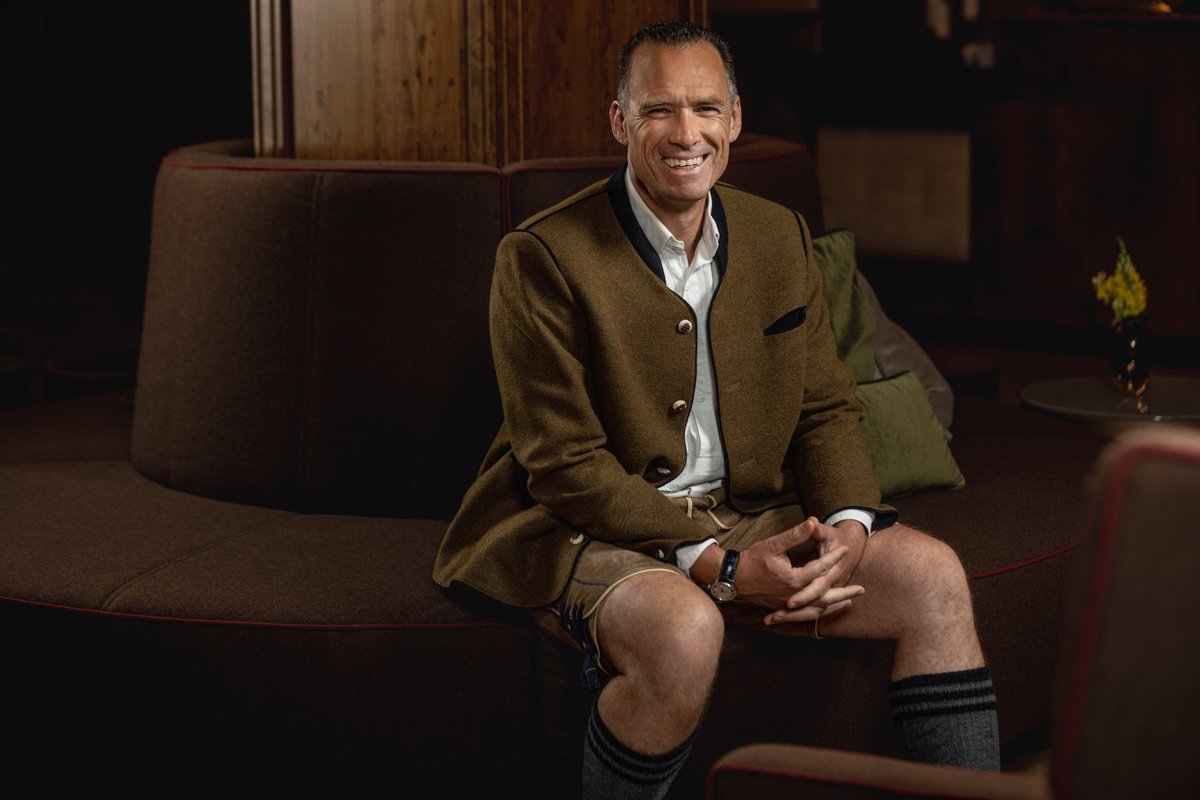 Lachender Mann in Trachtenjanker und Lederhose sitzt auf einem Rundsofa in einer dunklen, warmen Lounge