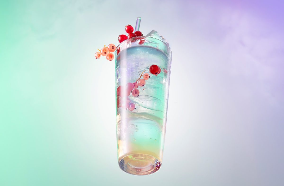 Schwebender Becher mit schimmerndem Drink, Eis und Johannisbeeren vor mint-fliederfarbenem Verlauf