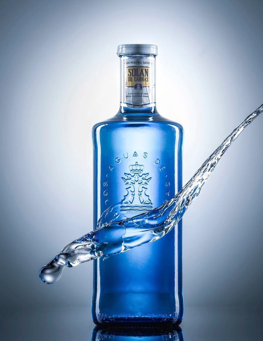 Blaue Glasflasche mit Wasserstrahl vor grau-blauem Studiohintergrund