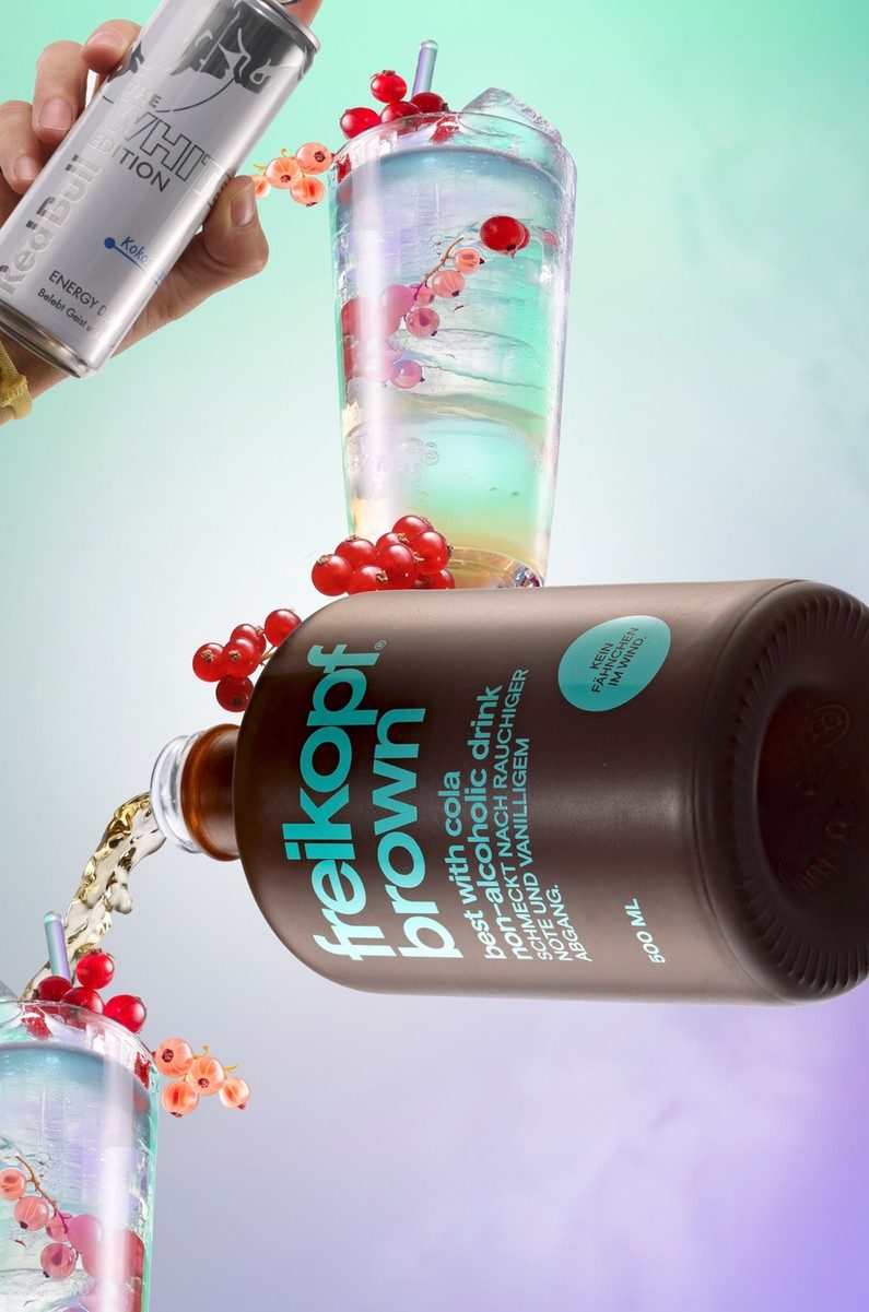 Schwebende braune Getränkeflasche mit Bechern, Eis und Johannisbeeren vor mint-fliederfarbenem Verlauf