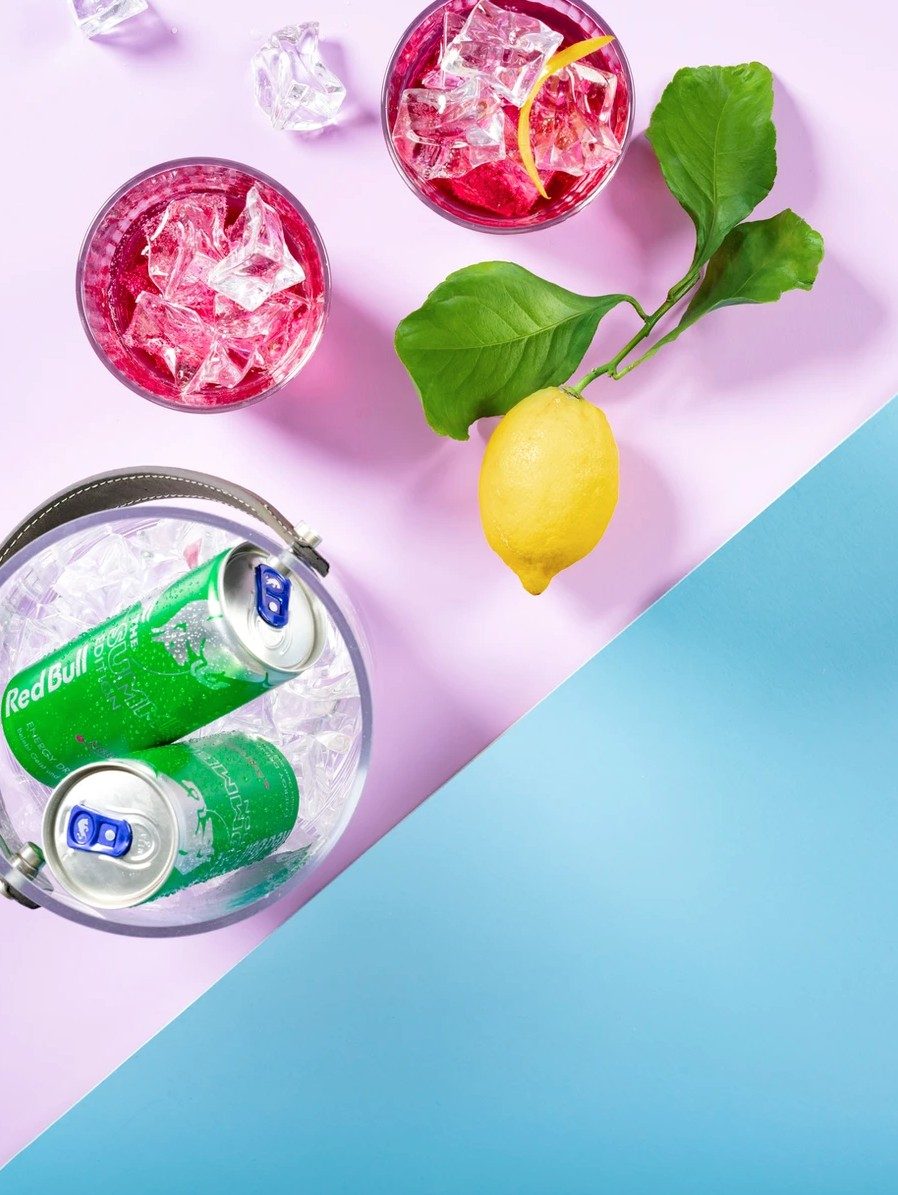 Eiskübel mit grünen Dosen, pinke Drinks und Zitrone von oben auf flieder-blauem Hintergrund