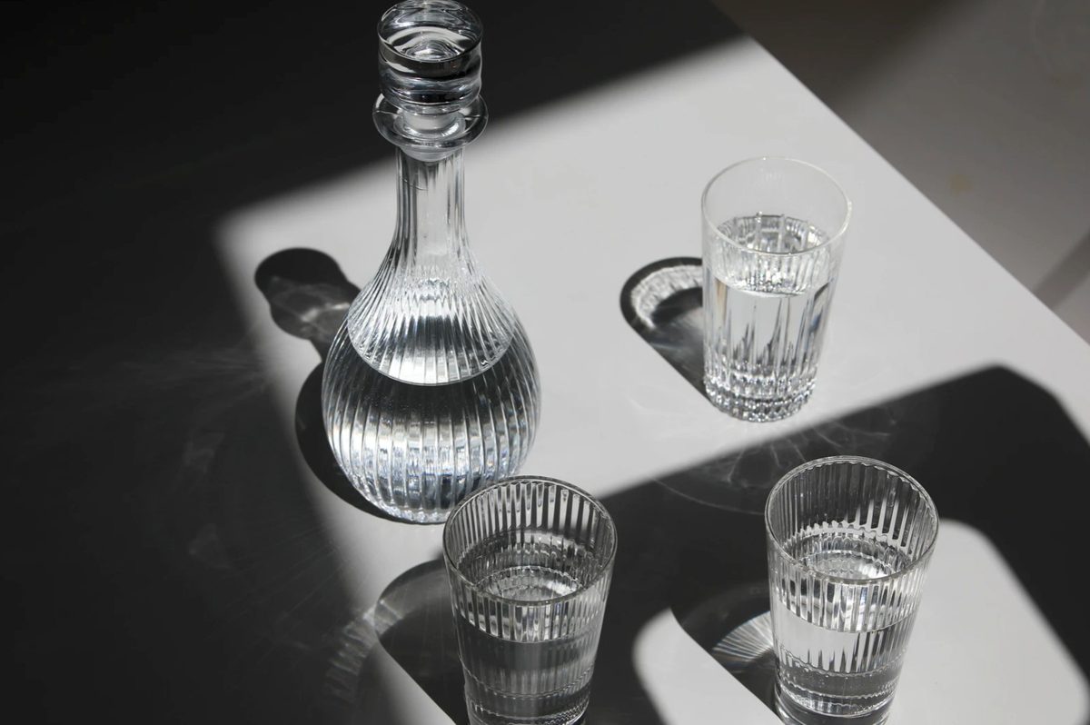 Geriffelte Glaskaraffe und Wassergläser mit grafischen Schlagschatten auf weißer Fläche
