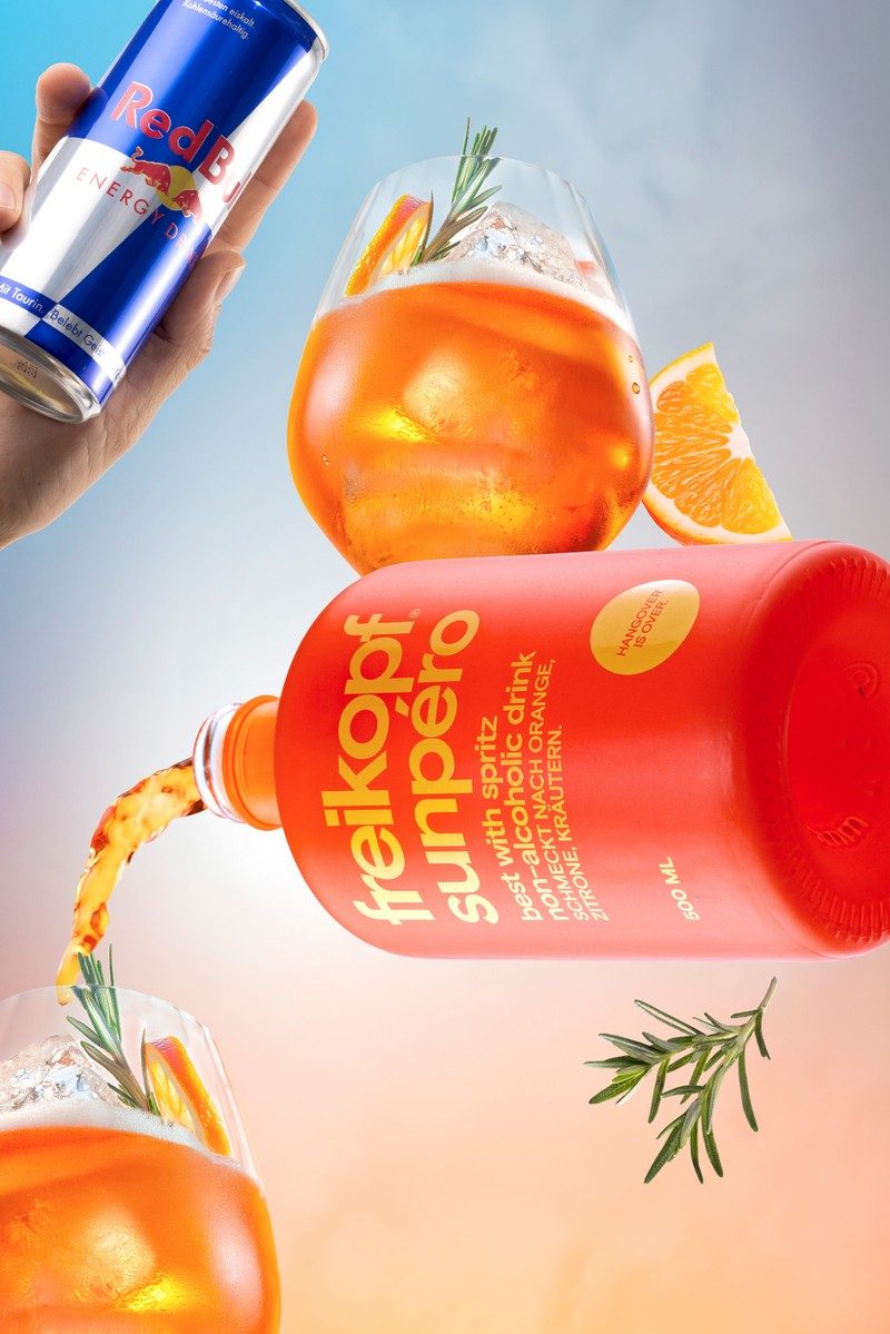 Schwebende orangefarbene Flasche mit Spritz-Glas, Orangenscheiben und Rosmarin vor blau-pfirsichfarbenem Verlauf