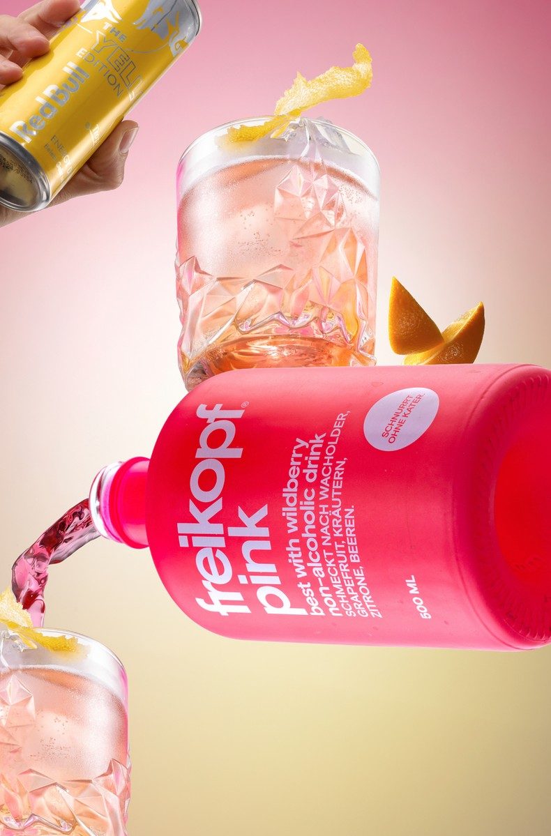 Schwebende pinke Flasche mit rosa Drinks, Eis und Zitrusfilets vor rosa-gelbem Verlauf