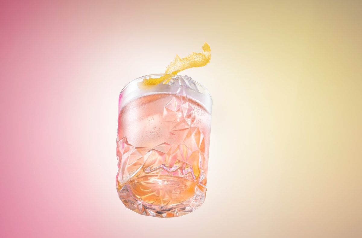 Schwebendes Kristallglas mit rosa Drink, Eis und Zitronenzeste vor pink-gelbem Verlauf