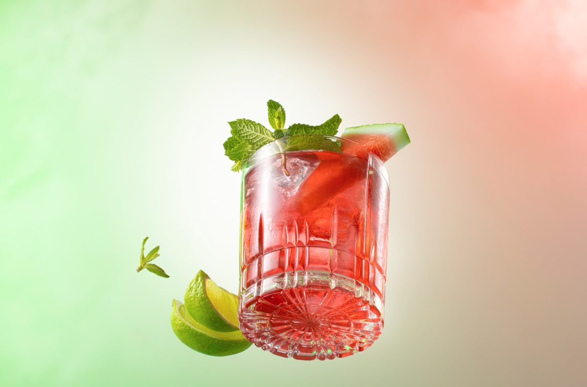 Schwebendes Glas mit rotem Drink, Minze, Melone und Limette vor grün-rosa Verlauf