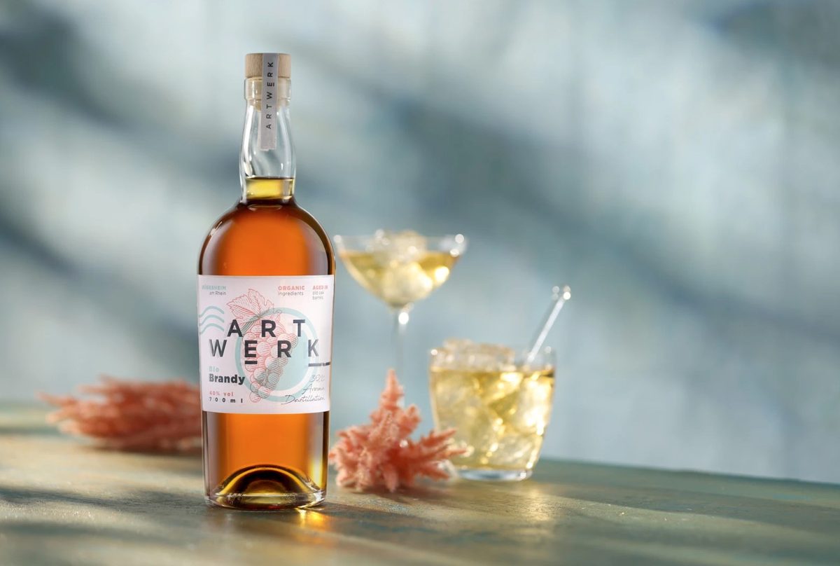 Spirituosenflasche mit zwei Gläsern und Korallen auf Holzfläche vor graublauem Hintergrund