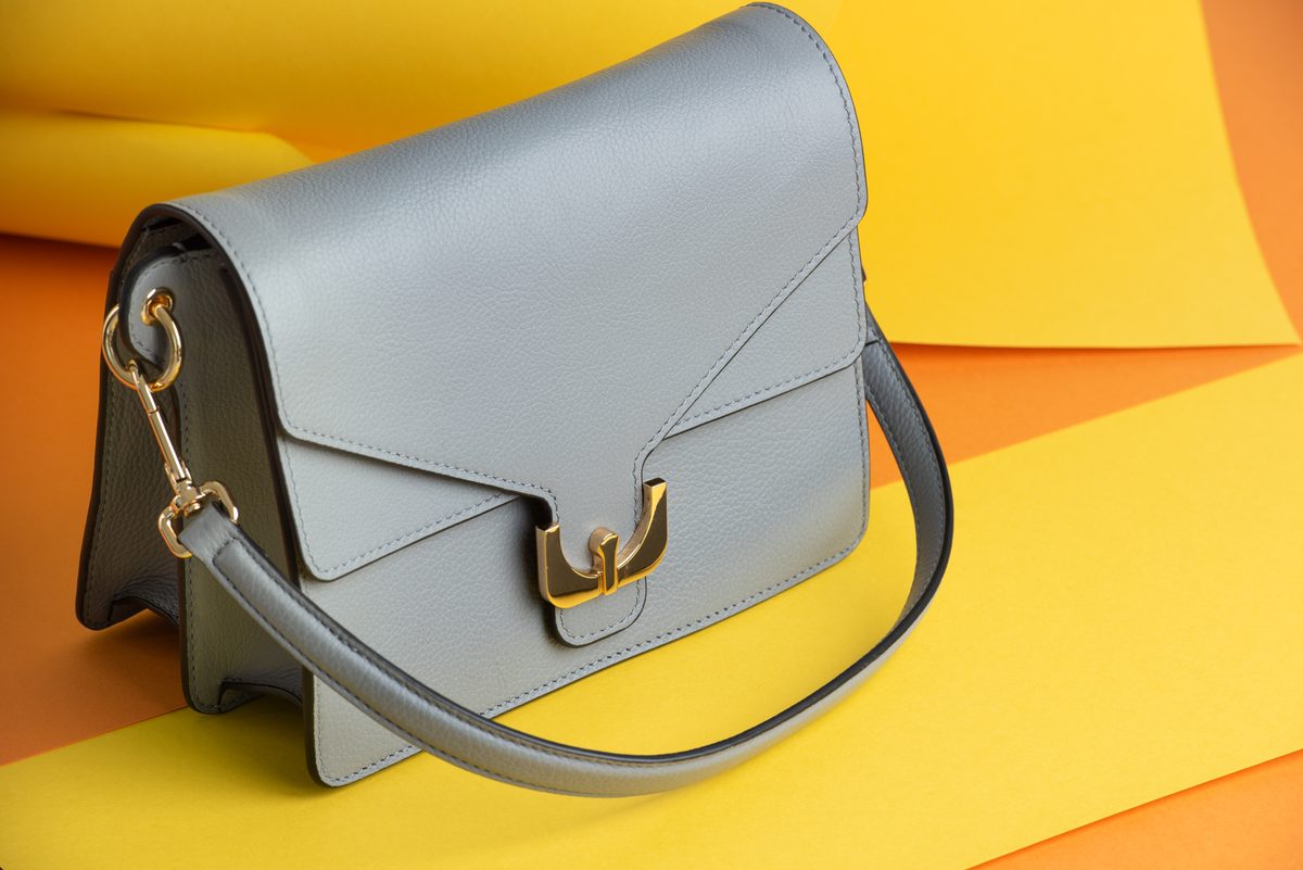 Hellgraue Ledertasche mit goldenem Verschluss vor gelbem und orangem Papierhintergrund
