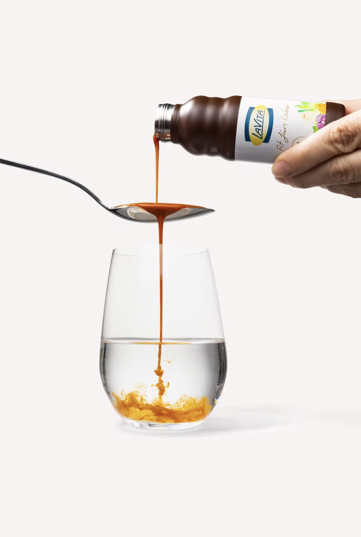 Flüssiges Konzentrat läuft von einem Löffel in ein Wasserglas, gehalten aus der LaVita Flasche