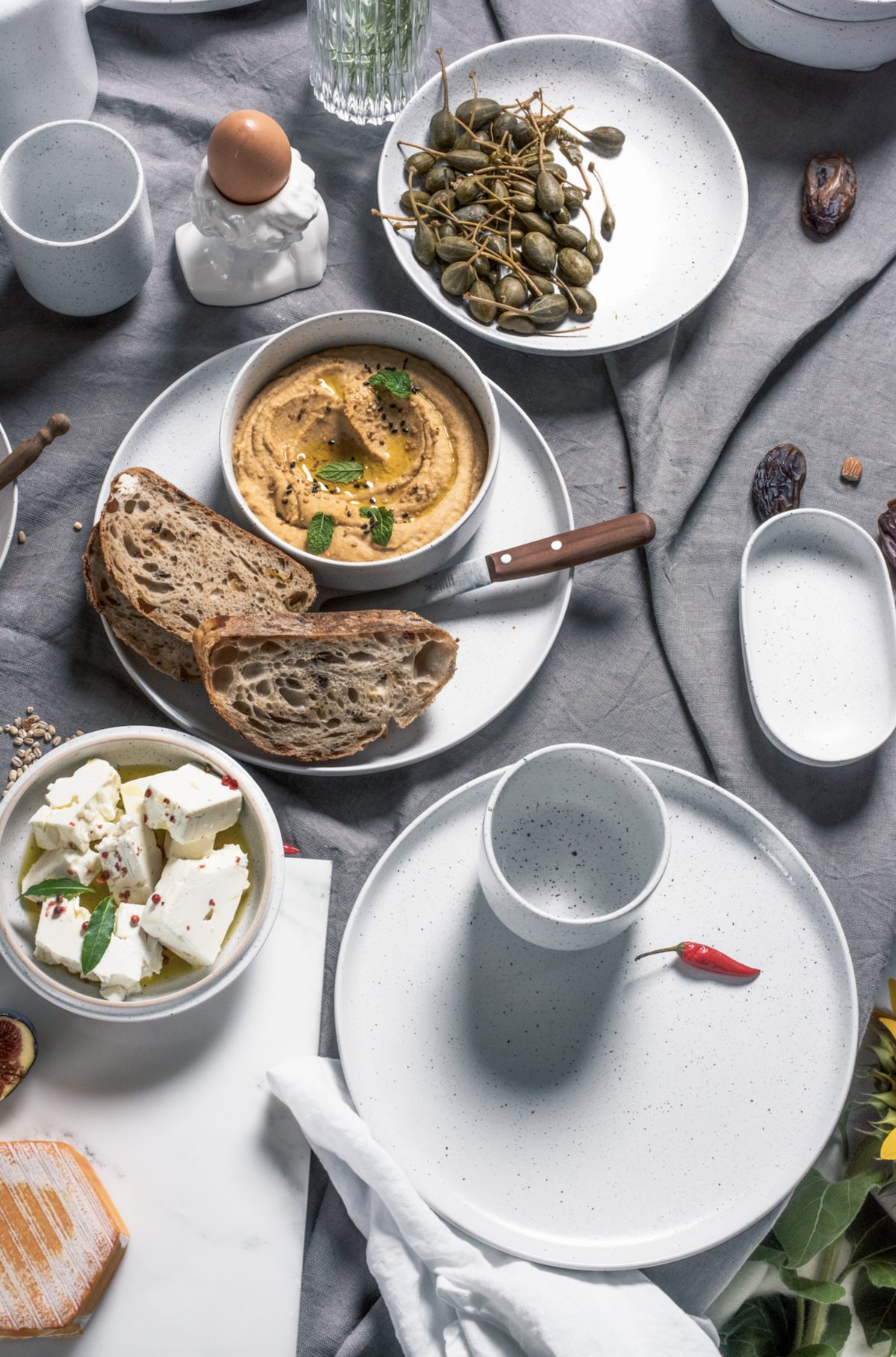 Aufsicht auf gesprenkeltes weißes Geschirr mit Hummus, Brot, Feta und Kapern auf grauem Leinen