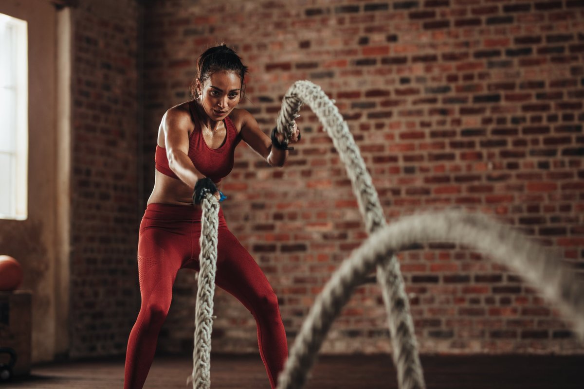 Frau in rotem Sportoutfit trainiert frontal mit zwei Battle Ropes vor einer Backsteinwand