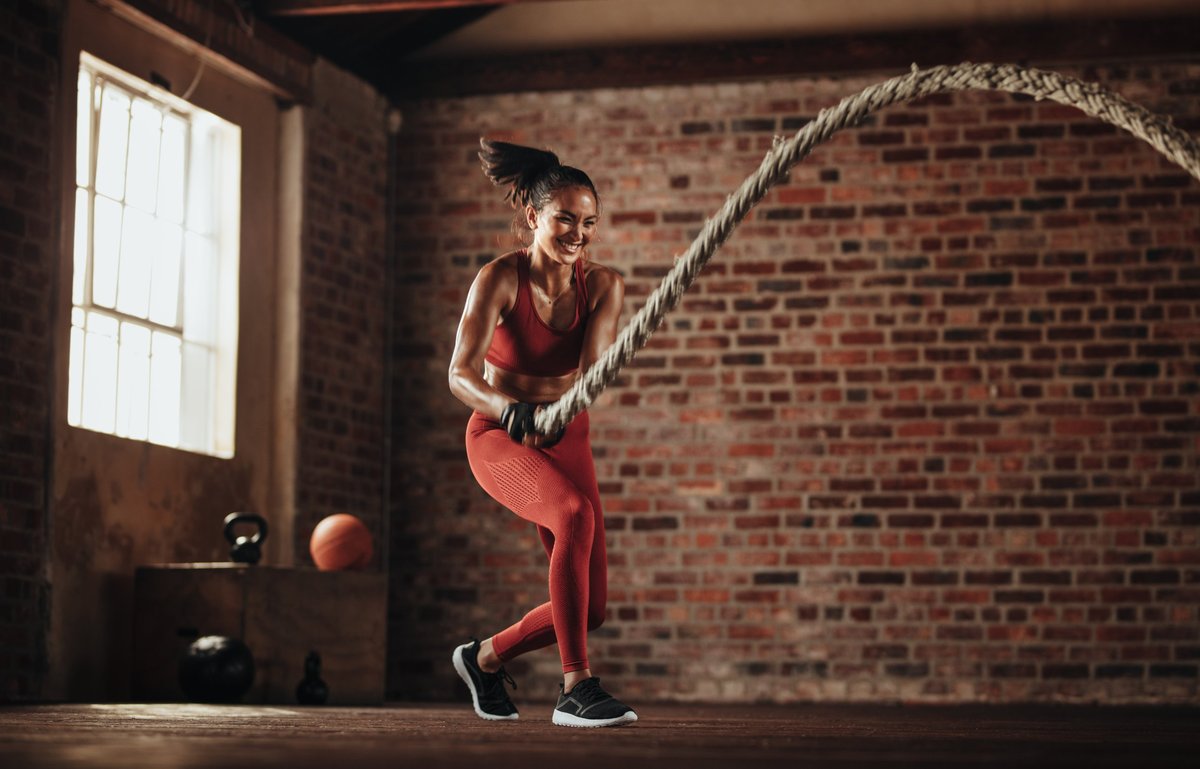 Lachende Frau in rotem Sportoutfit trainiert mit Battle Ropes in einem Loft mit Backsteinwand